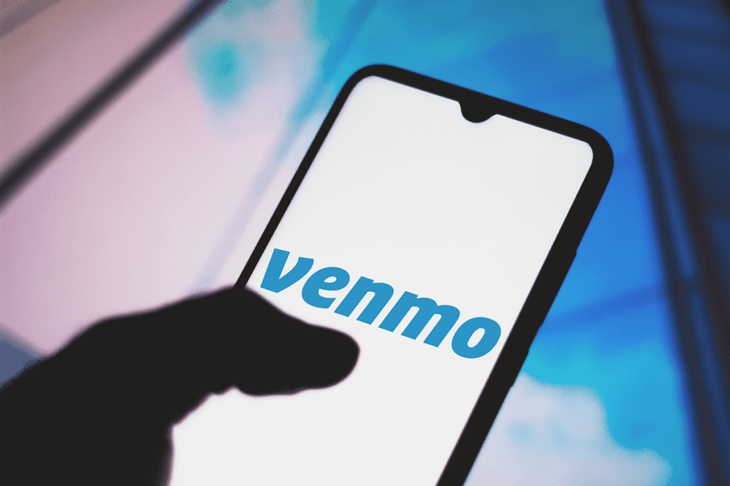 Image shows a silouetted hand holding a phone, with their thumb over the Venmo logo in blue letters illuminated over a white screen