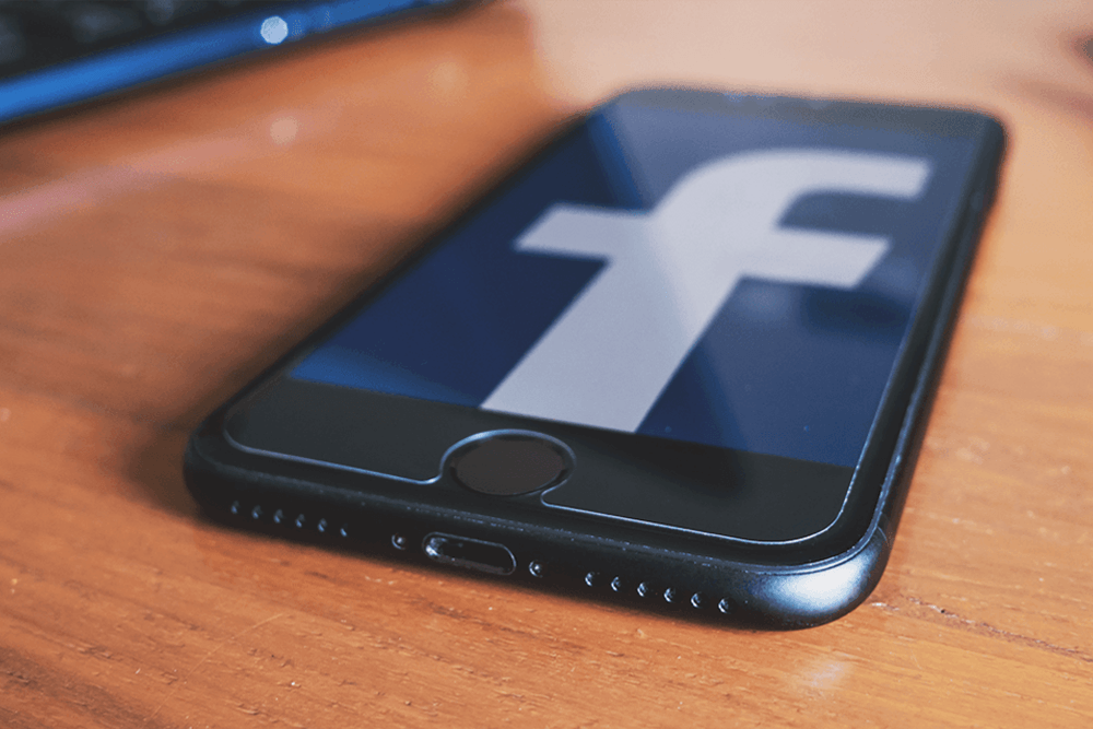 A smartphone microphone is in focus on a screen displaying the Facebook logo, implying listening