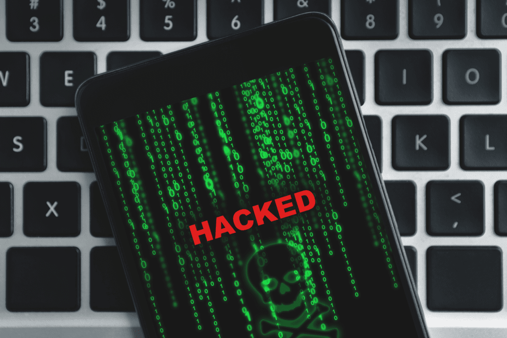 A mobile phone rests on a laptop keyboard as its display shows the word "Hacked" in red letters with a binary skull behind it. How to know if your phone is hacked visualized