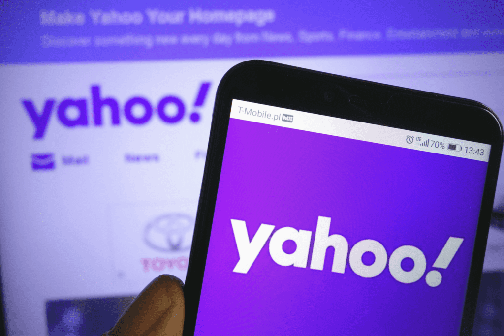 A phone with the Yahoo company logo is held in front of a computer monitor displaying the Yahoo homepage while the user determines how to delete a Yahoo account.