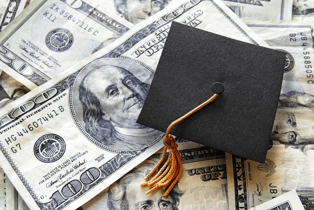 A small graduation cap rests on a pile of $100 bills to symbolize student loans and the student loan relief scams people face.