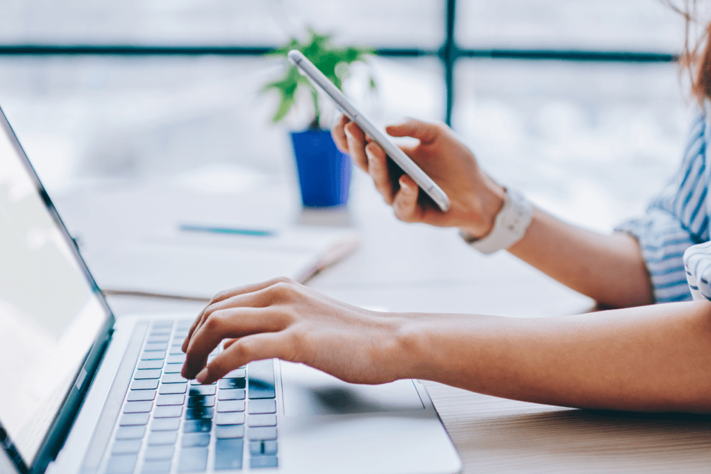 A person uses one hand to type on a laptop and the other to type on a phone, visualizing choosing between an app vs website of an online service