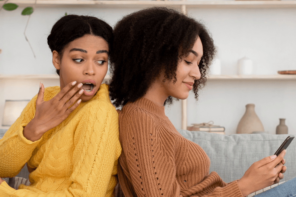 Shoulder surfing example with two women sit on the couch with one peeking over the shoulder to view the phone activity of the other